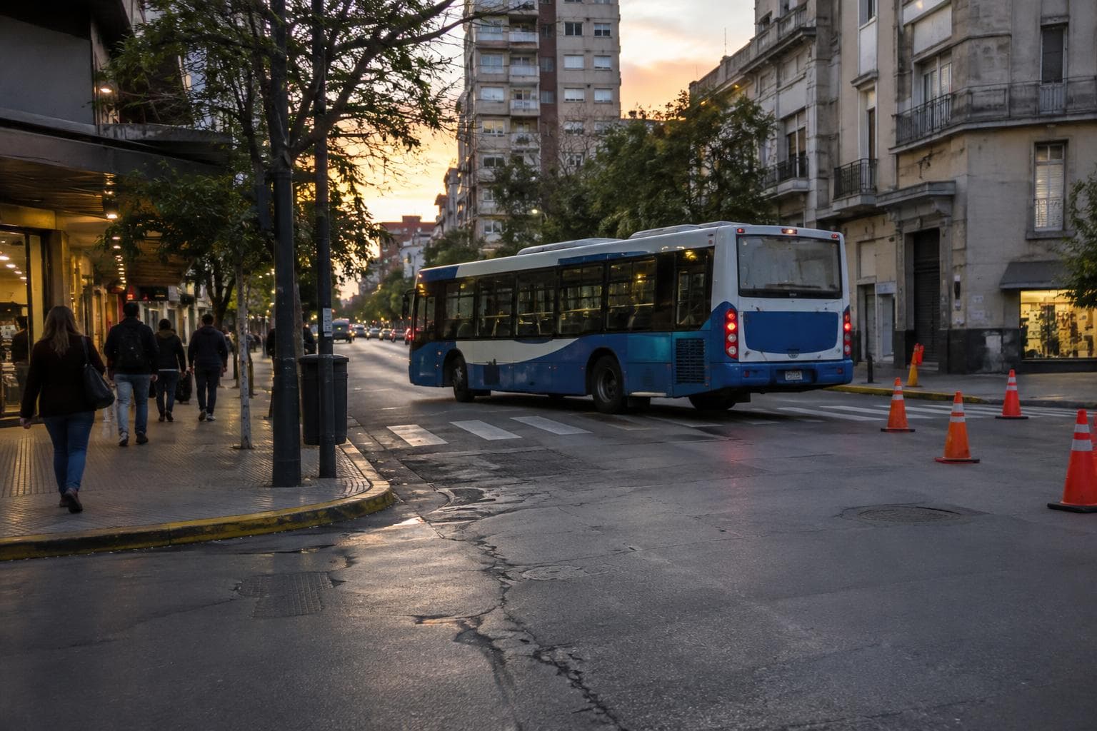 An urban Rosario street at dusk, with pedestrians, traffic cones and a bus taking a detour.