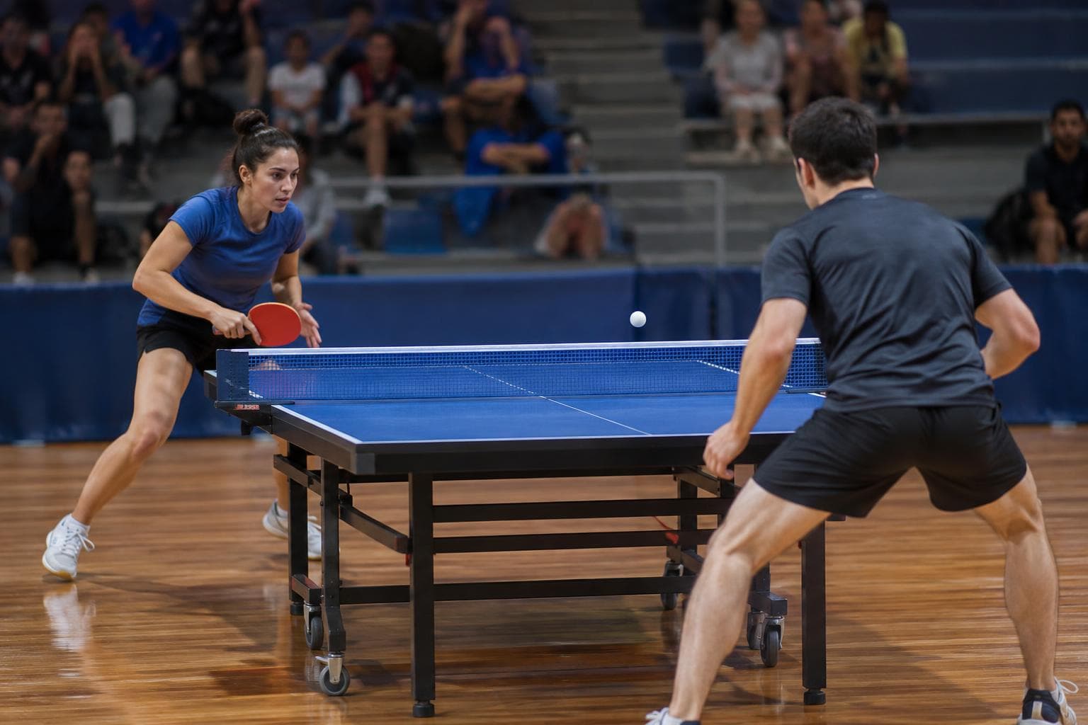 Un partido de tenis de mesa en un polideportivo, con jugadores y público desenfocado.