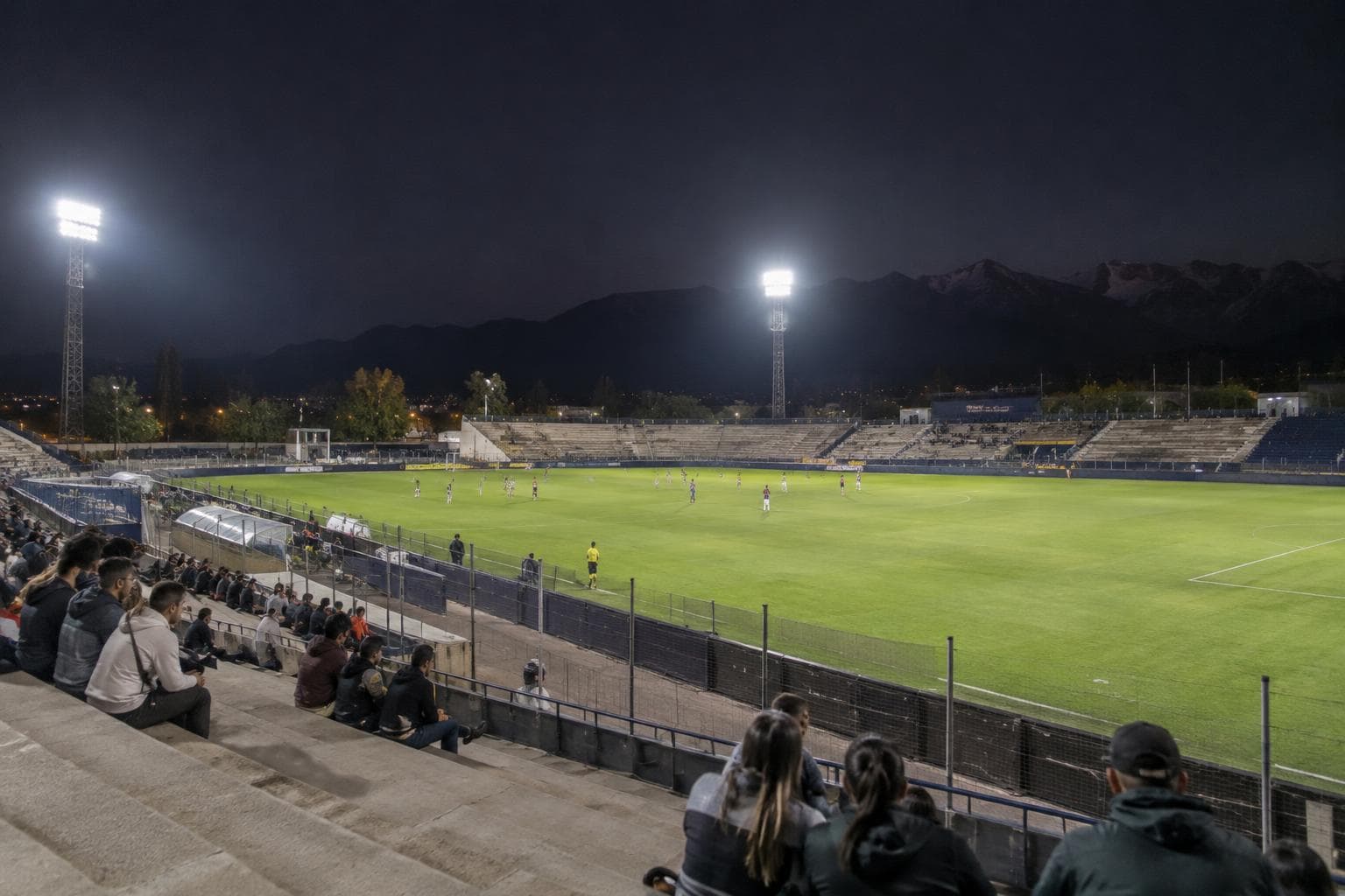 De carácter ilustrativo, de un estadio de fútbol nocturno en Mendoza con jugadores genéricos a lo lejos y tribunas iluminadas.