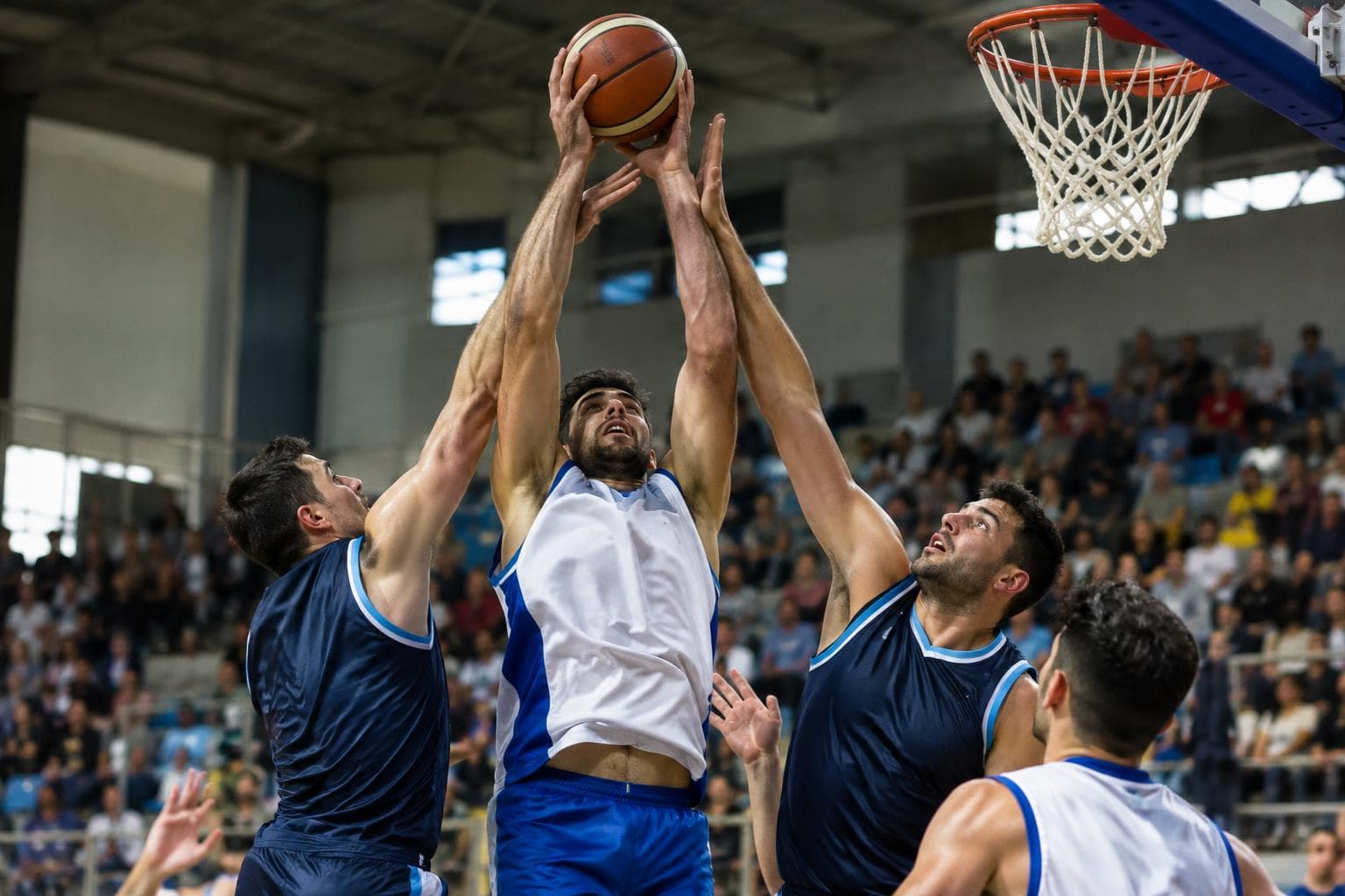 Basketball players contesting a rebound in an indoor arena, with spectators blurred in the background.